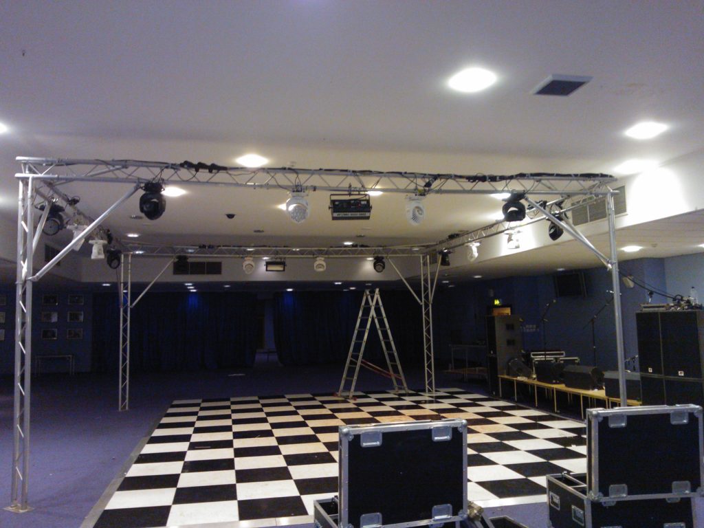 Photo taken during set up (room lights on) shows truss rig with event lighting with flight cases in the foreground in an otherwise empty room