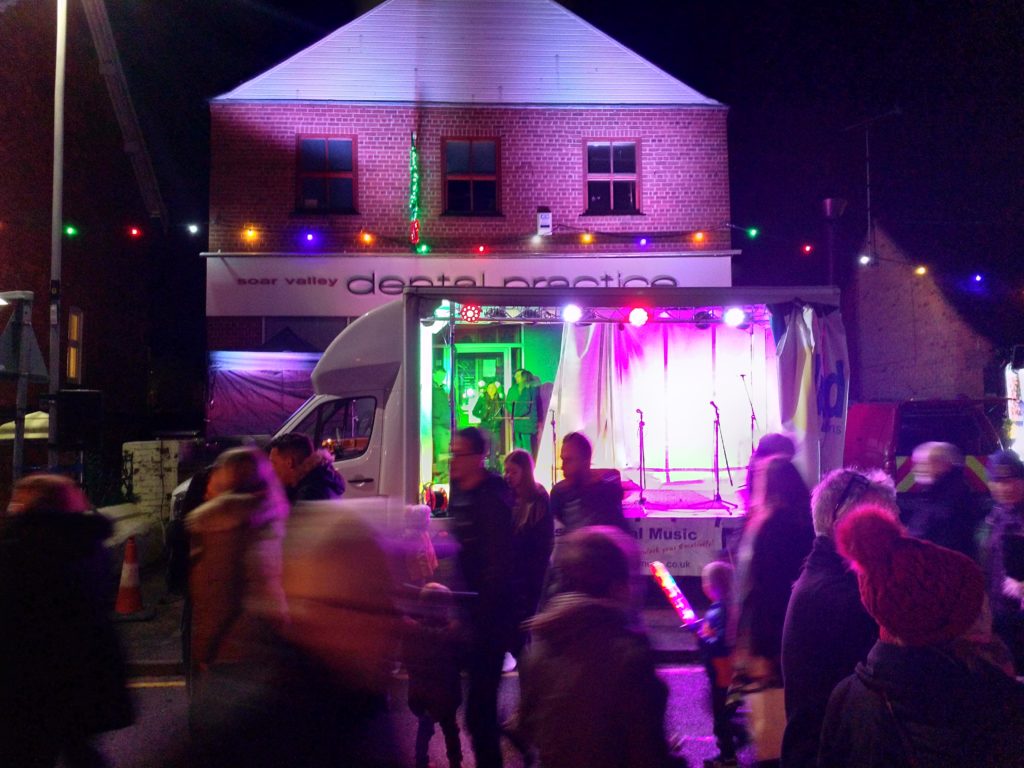Photo shows the stage lit up between acts at the Barrow Upon Soar Christmas Lights switch on.

The stage and building behind are washed in purple, and the backstage area contrasts in green. We didn't pick the colours(!)